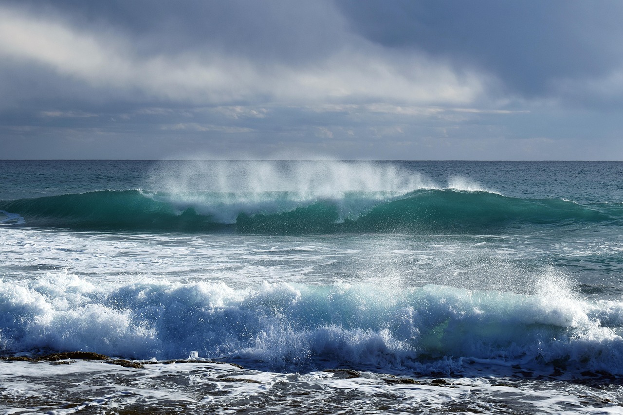 A teal wave breaking with spray under a heavy grey sky